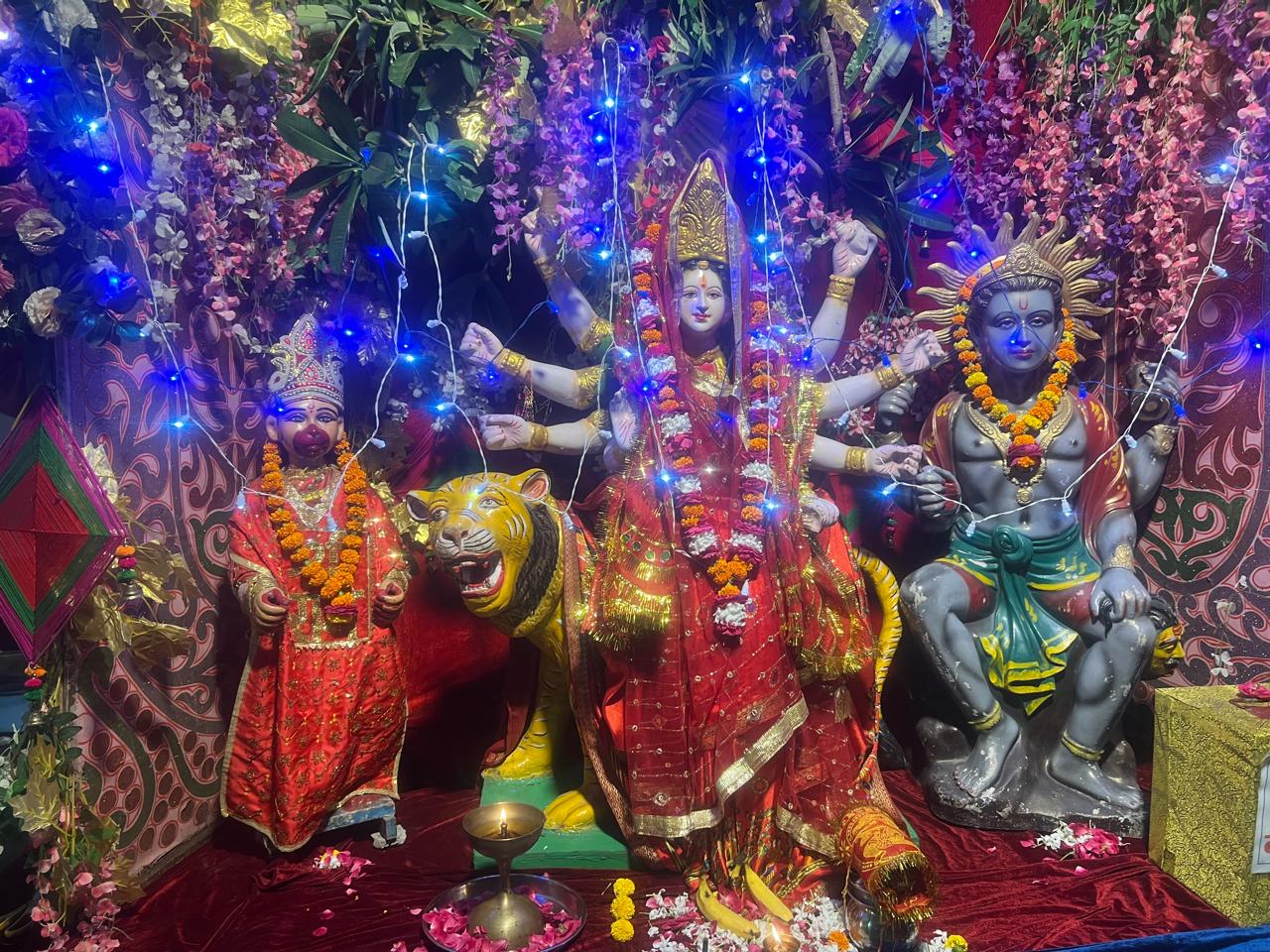 A vibrant display of Hindu deities, featuring a multi-armed goddess on a tiger, adorned with lights and floral decorations, reminiscent of a जागरण celebration, accompanied by two other deities in traditional attire.